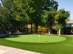 distant view of a backyard putting green kit. There are 3 holes, grass, brick, and trees surrounding the beautiful turf.