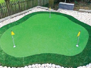 outdoor putting green kit with 3 holes and pins. it has artificial grass turf, fringe, and small rock landscaping in the backyard with a fence surrounding it