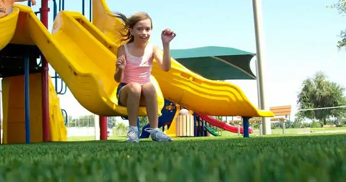 child sliding down slide and looking at camera with close up of artificial grass turf installation