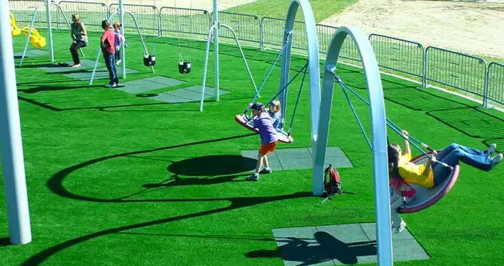2 children swinging in a swing set area installed on top of artificial grass