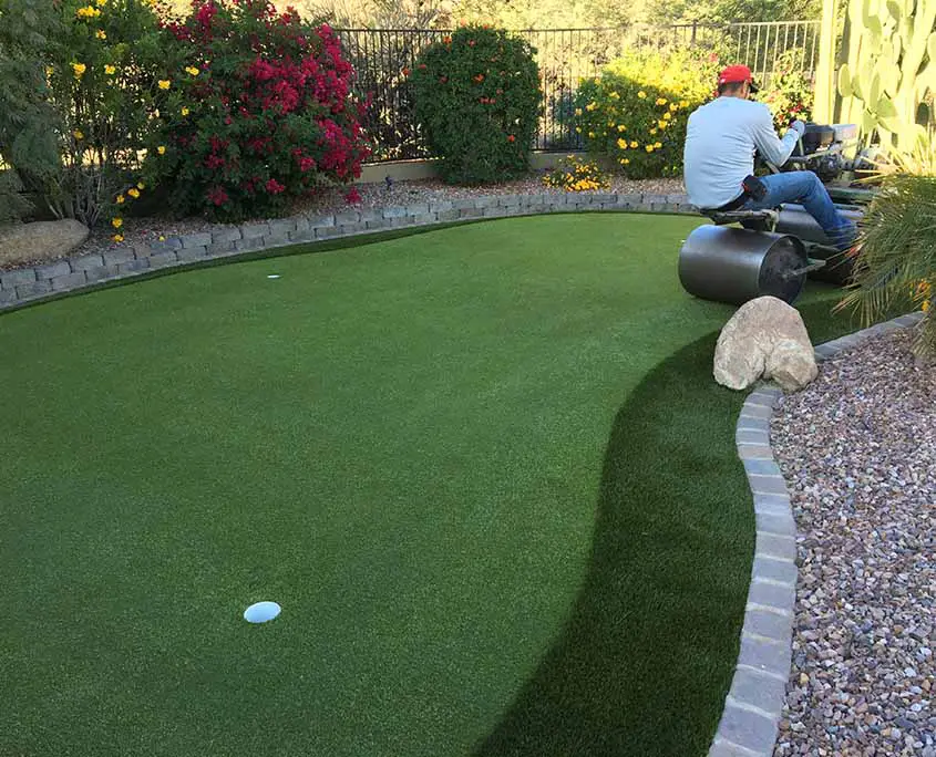 artificial grass installer driving a turf compactor over a backyard putting green. there are flowers and rock landscaping surrounding the course.