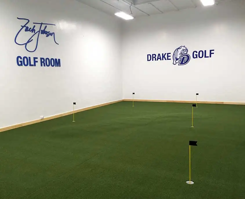 indoor artificial grass putting green installation at drake university in des moines, iowa. there are 5 pins and holes and the course is flat. the wall contains artwork with a logo saying "zach johnson golf room" and another logo saying "drake golf"