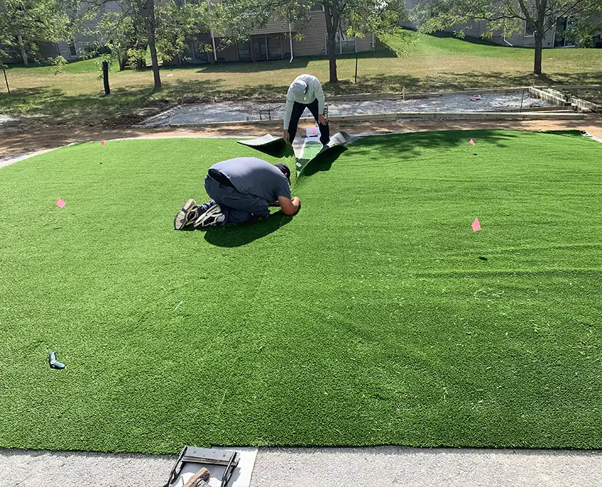 2 artificial turf installers in des moines, iowa. 1 is on his knees lining up the turf. the other is laying the turf. it is a residential installation in a backyard.