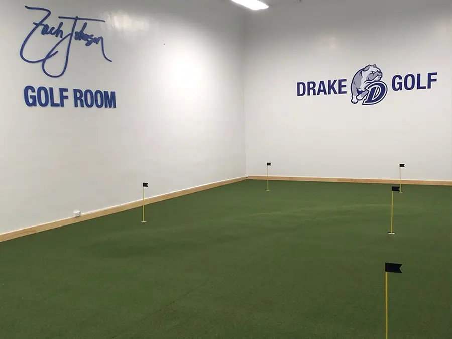 indoor putting green using artificial grass at drake university in des moines, iowa. the wall has signage that reads "zach johnson golf room" and "drake golf"